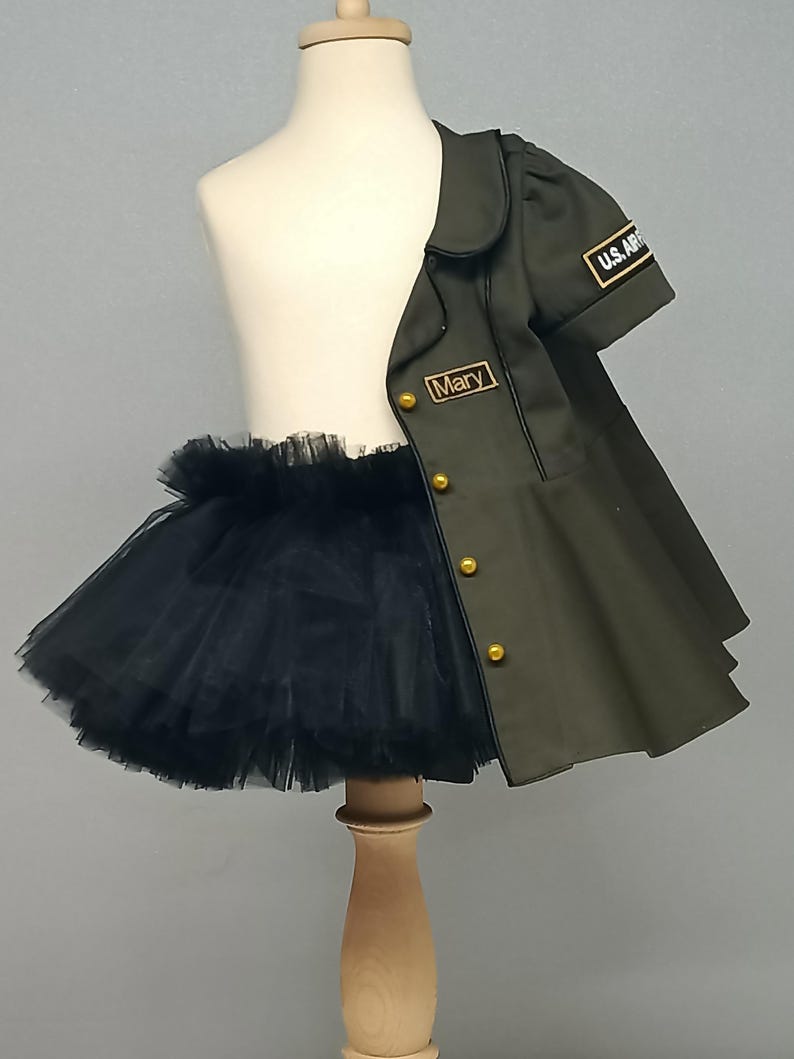 May include: A child's olive green jacket with gold buttons and "U.S. ARMY" patch, paired with a black tulle tutu. The jacket has the name "Mary" embroidered on it. The outfit is displayed on a mannequin.
