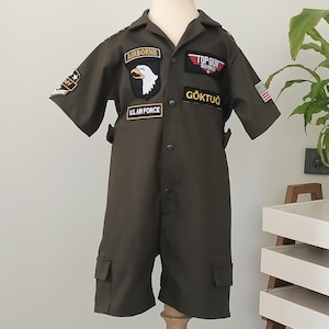 Puede incluir: Mono verde oliva con parches, incluyendo "AIRBORNE", "TOP GUN MAVERICK" y "U.S. AIR FORCE". El mono de manga corta tiene dos bolsillos delanteros y el texto "G&Ouml;KTUĞ". Se muestra en un maniqu&iacute;.