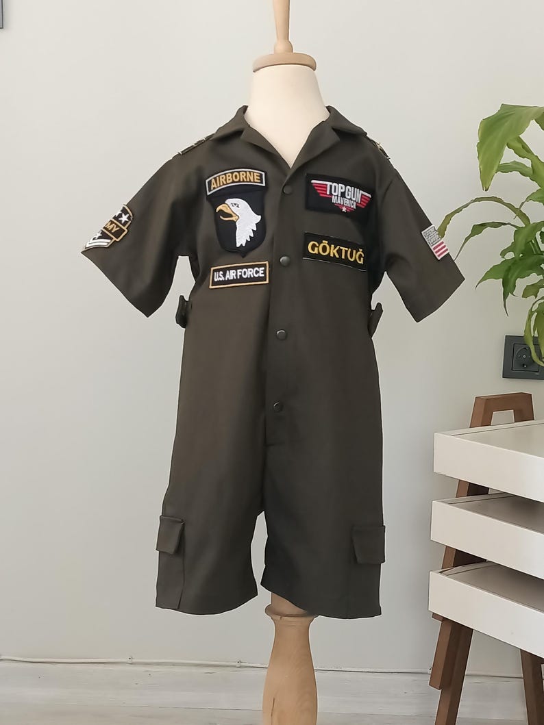 Puede incluir: Mono verde oliva con parches, incluyendo "AIRBORNE", "TOP GUN MAVERICK" y "U.S. AIR FORCE". El mono de manga corta tiene dos bolsillos delanteros y el texto "G&Ouml;KTUĞ". Se muestra en un maniqu&iacute;.