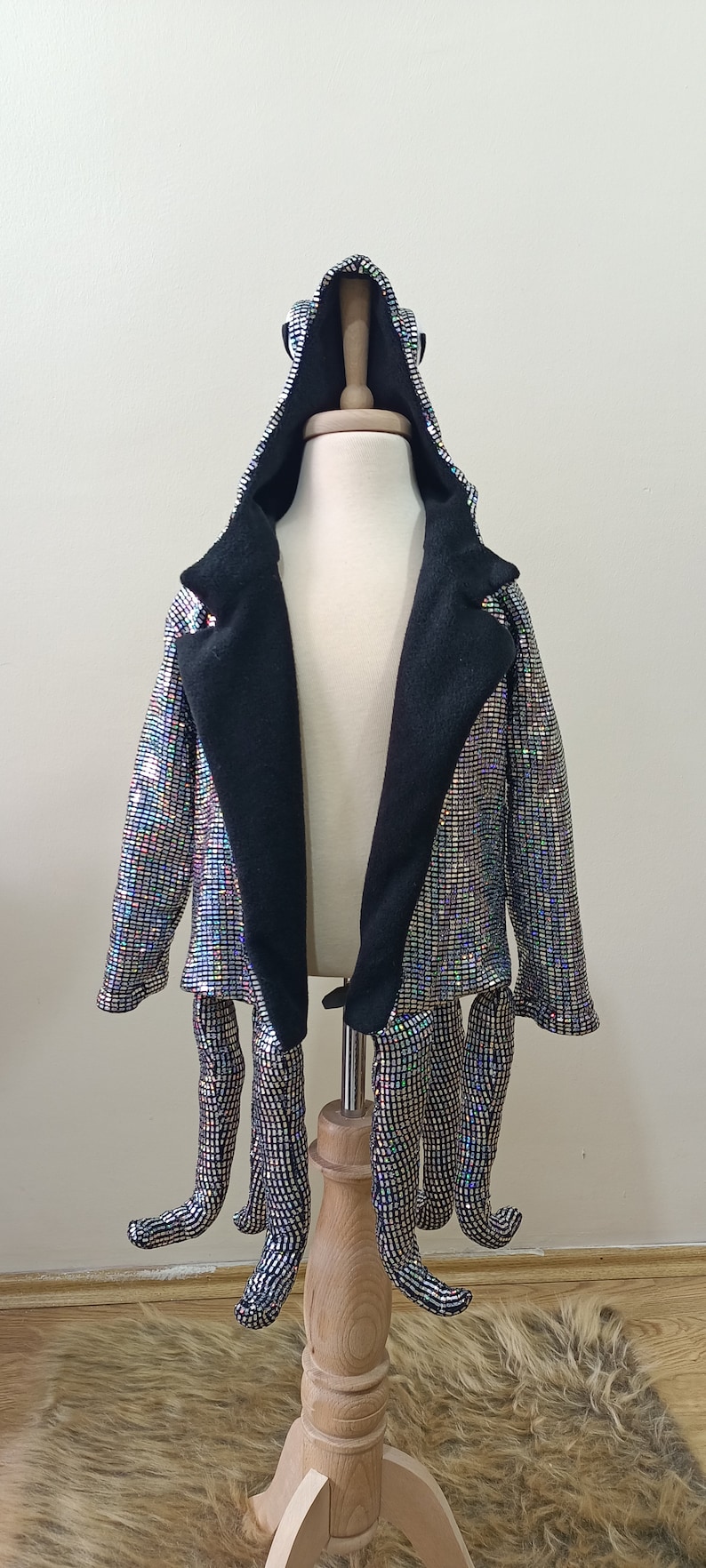 Puede incluir: Una chaqueta con capucha de lentejuelas plateadas con forro negro y ocho largas tentaculos de lentejuelas plateadas unidas a la parte inferior. La chaqueta est&aacute; en un maniqu&iacute; de madera.