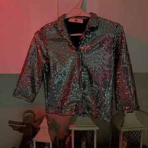 May include: A silver sequined jacket with a black collar and cuffs hangs on a white hanger. The jacket has a grid pattern of small, reflective sequins. The sleeves are three-quarter length. The background includes decorative lanterns and a shelf.