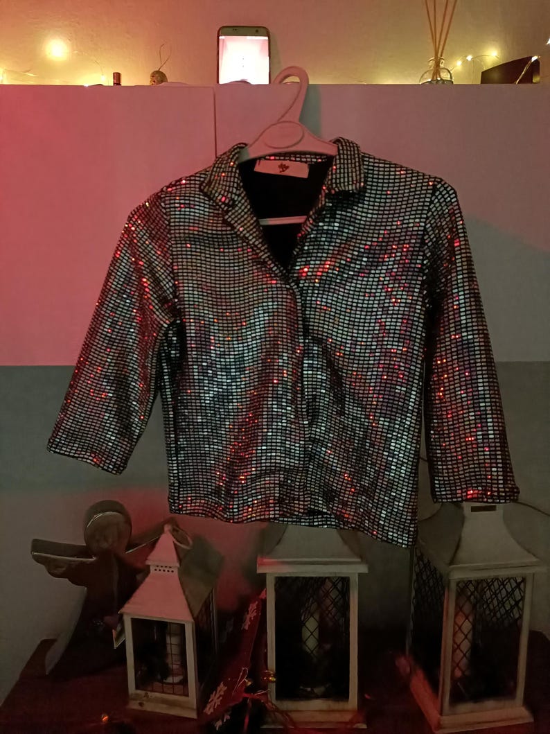 May include: A silver sequined jacket with a black collar and cuffs hangs on a white hanger. The jacket has a grid pattern of small, reflective sequins. The sleeves are three-quarter length. The background includes decorative lanterns and a shelf.