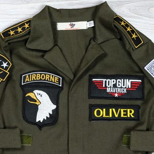 May include: Olive green Top Gun Maverick flight suit with patches. Features an "AIRBORNE" patch with an eagle, a "TOP GUN MAVERICK" patch, and a name patch that says "OLIVER". Includes gold star epaulettes and a small label that says "Fashion Teenagers".