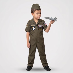 May include: A child in an olive green Top Gun flight suit costume, complete with a matching cap. The costume features patches, including an Airborne patch with a white eagle and the name "Oliver". The child is holding a gray toy airplane.