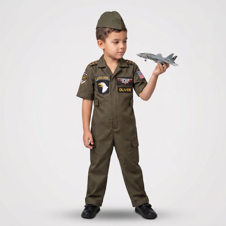 May include: A child in an olive green Top Gun flight suit costume, complete with a matching cap. The costume features patches, including an Airborne patch with a white eagle and the name "Oliver". The child is holding a gray toy airplane.