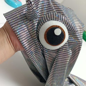 K&ouml;nnte beinhalten: Ein handgefertigtes Kost&uuml;mteil aus silbernem Stoff mit einem Gittermuster. Es hat ein gro&szlig;es Filzauge mit brauner Iris und schwarzer Pupille sowie ein kleines Horn. Der Stoff ist reflektierend.