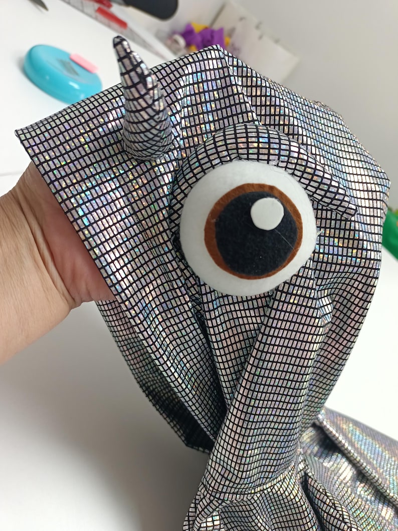 K&ouml;nnte beinhalten: Ein handgefertigtes Kost&uuml;mteil aus silbernem Stoff mit einem Gittermuster. Es hat ein gro&szlig;es Filzauge mit brauner Iris und schwarzer Pupille sowie ein kleines Horn. Der Stoff ist reflektierend.