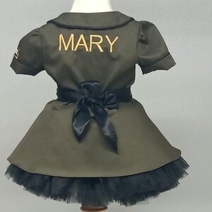 May include: Olive green dress with a black tulle skirt and a black bow at the waist. The word "MARY" is embroidered in gold on the back of the dress. The dress has short sleeves and a collared neckline. A small patch is visible on the left sleeve.