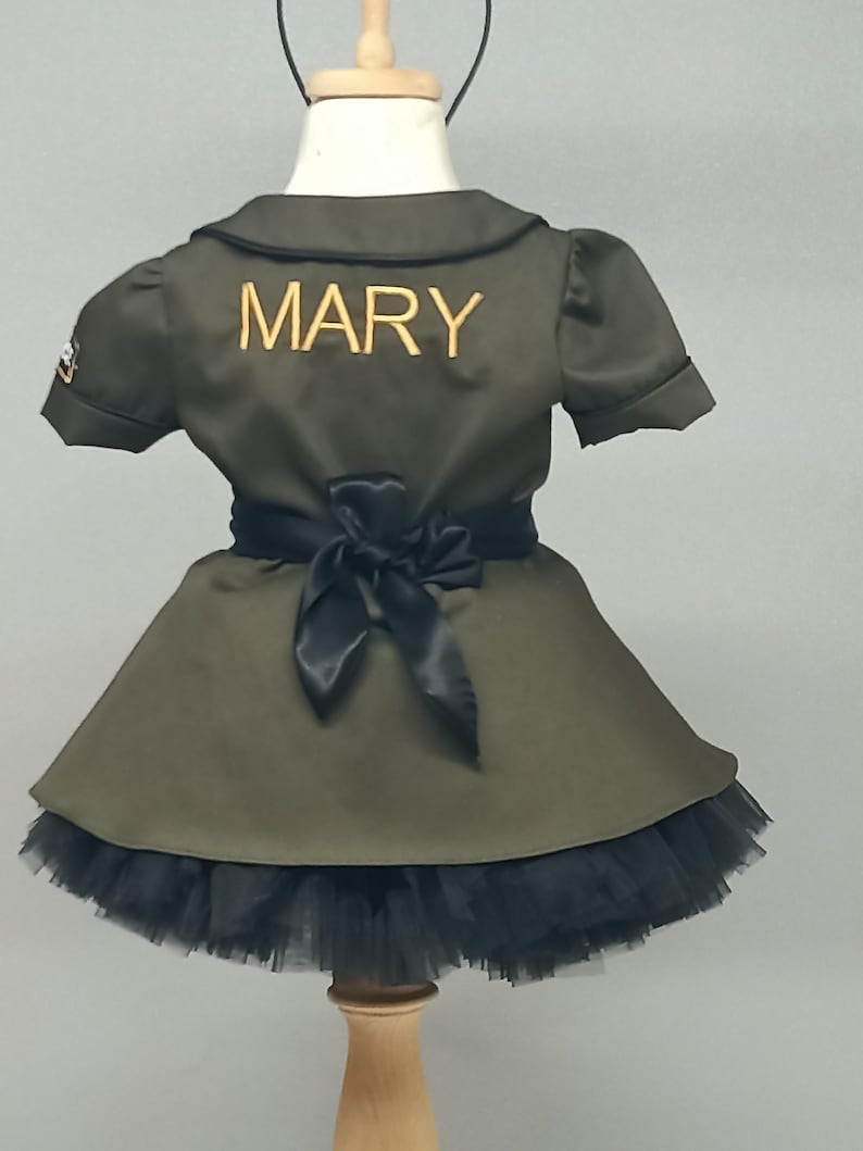 May include: Olive green dress with a black tulle skirt and a black bow at the waist. The word "MARY" is embroidered in gold on the back of the dress. The dress has short sleeves and a collared neckline. A small patch is visible on the left sleeve.