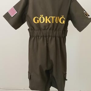 Puede incluir: Mono verde oliva oscuro con mangas cortas, cuello camisero y cintura el&aacute;stica. Cuenta con un parche de la bandera estadounidense en la manga izquierda y la palabra "G&Ouml;KTUĞ" bordada en amarillo en la espalda. Incluye dos bolsillos de carga.