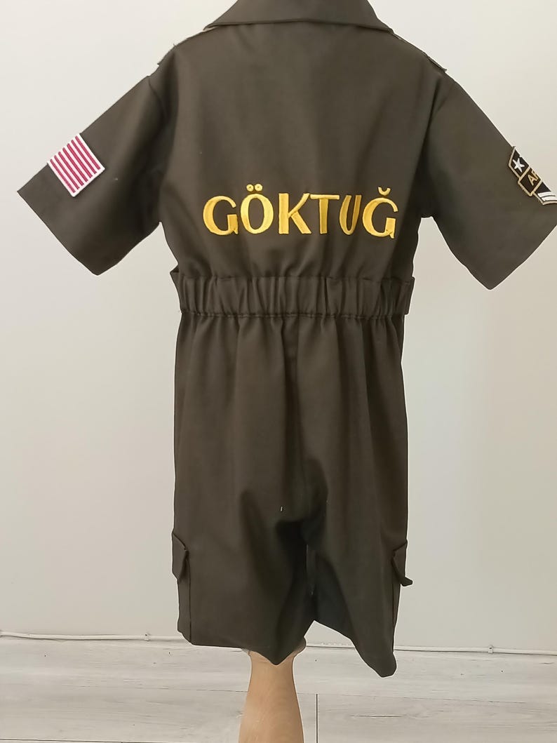 Puede incluir: Mono verde oliva oscuro con mangas cortas, cuello camisero y cintura el&aacute;stica. Cuenta con un parche de la bandera estadounidense en la manga izquierda y la palabra "G&Ouml;KTUĞ" bordada en amarillo en la espalda. Incluye dos bolsillos de carga.