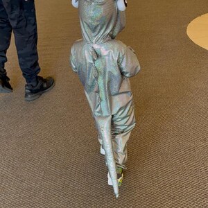 K&ouml;nnte beinhalten: Ein Kind in einem silbernen, schillernden Drachenkost&uuml;m mit Kapuze und Schwanz, das einen Flur entlanggeht. Das Kost&uuml;m hat gro&szlig;e, runde Augen mit wei&szlig;em Rand auf der Kapuze. Das Kind tr&auml;gt wei&szlig;e Schuhe mit gr&uuml;nen Akzenten.