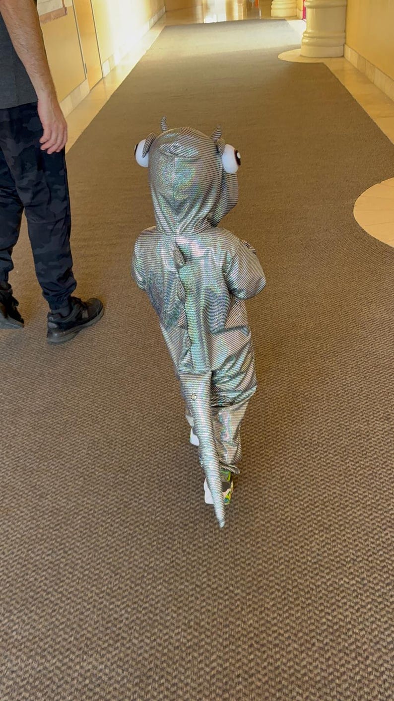 K&ouml;nnte beinhalten: Ein Kind in einem silbernen, schillernden Drachenkost&uuml;m mit Kapuze und Schwanz, das einen Flur entlanggeht. Das Kost&uuml;m hat gro&szlig;e, runde Augen mit wei&szlig;em Rand auf der Kapuze. Das Kind tr&auml;gt wei&szlig;e Schuhe mit gr&uuml;nen Akzenten.