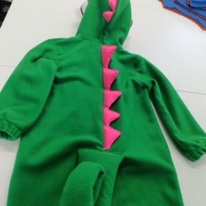 May include: A green dinosaur costume with a hood and attached tail. The costume features pink accents along the spine and on the hood. The long-sleeved jumpsuit is made of a soft material.