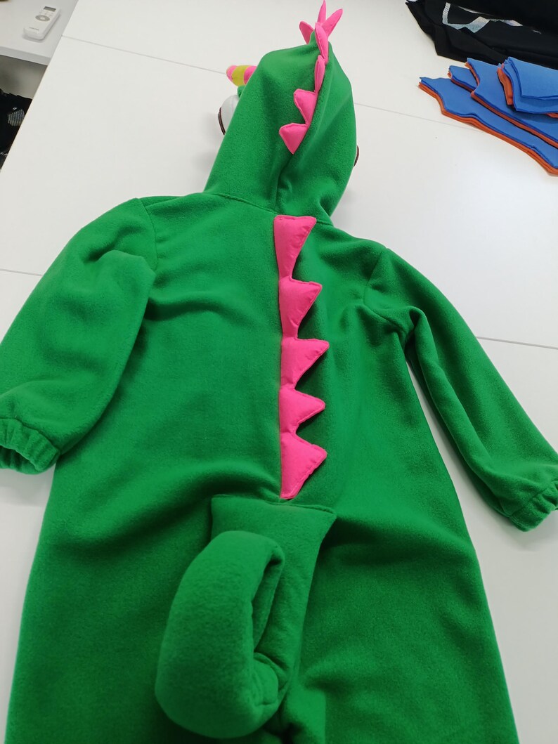 May include: A green dinosaur costume with a hood and attached tail. The costume features pink accents along the spine and on the hood. The long-sleeved jumpsuit is made of a soft material.