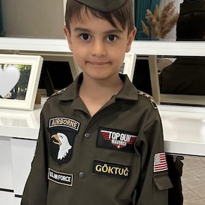 May include: A young person wearing an olive green Air Force uniform and cap. The uniform features patches that read "AIRBORNE", "TOP GUN MAVERICK", and "US AIR FORCE". The name "G&Ouml;KTUĞ" is also displayed on the uniform.