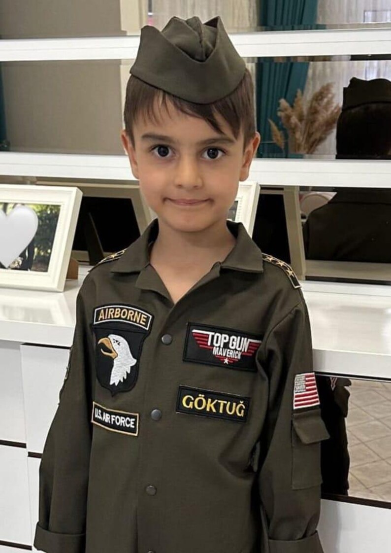 May include: A young person wearing an olive green Air Force uniform and cap. The uniform features patches that read "AIRBORNE", "TOP GUN MAVERICK", and "US AIR FORCE". The name "G&Ouml;KTUĞ" is also displayed on the uniform.
