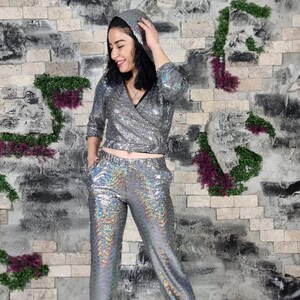 Puede incluir: Un pantal&oacute;n de vestir plateado con lentejuelas y una parte superior con capucha. El pantal&oacute;n de vestir tiene un brillo iridiscente y lo lleva puesto una persona de pie frente a una pared de ladrillo.