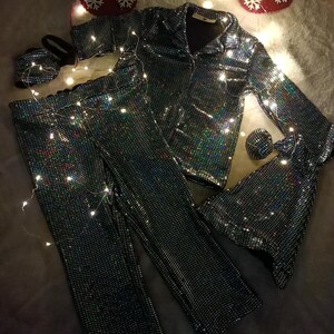 May include: A matching pajama set with a shimmering, iridescent finish. The set includes a button-down shirt, pants, a sleep mask, and a scrunchie. Christmas stockings and fairy lights are in the background.