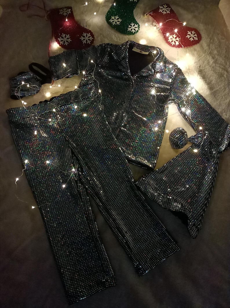 May include: A matching pajama set with a shimmering, iridescent finish. The set includes a button-down shirt, pants, a sleep mask, and a scrunchie. Christmas stockings and fairy lights are in the background.