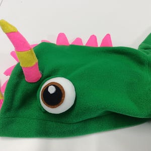 May include: A green fleece dragon hood with pink and yellow accents. The hood features a large, round eye and pink spikes along the back. The horns are pink with yellow stripes.