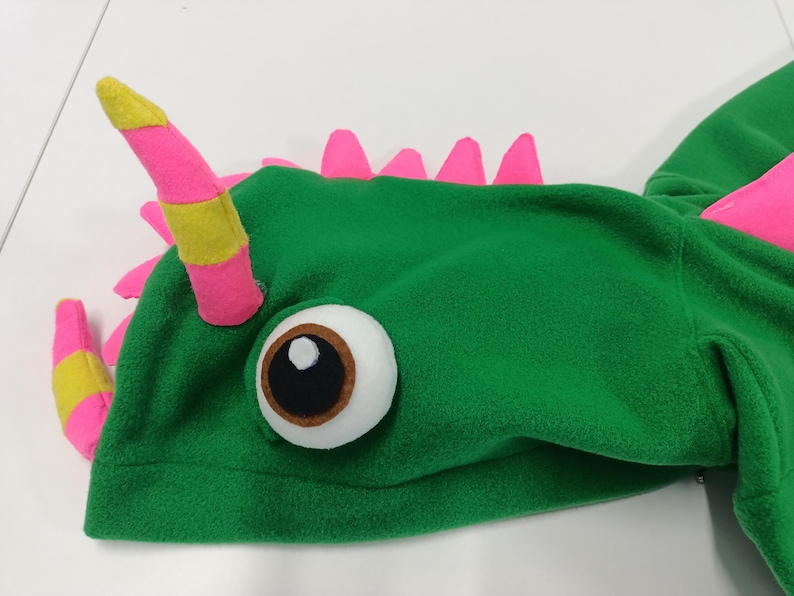 May include: A green fleece dragon hood with pink and yellow accents. The hood features a large, round eye and pink spikes along the back. The horns are pink with yellow stripes.
