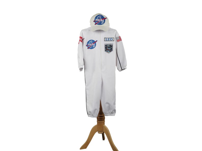 Może przedstawiać: Biały kostium astronauty z naszywkami NASA i pasującą czapką. Kostium ma czarne wykończenia i napis "LEO" na piersi. Czapka ma logo NASA. Kostium jest prezentowany na drewnianym stojaku.