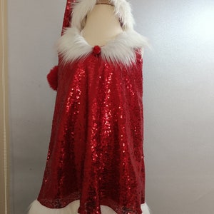 May include: A red sequined Santa dress with a white faux fur trim around the hood, neckline, and hem. The dress has a sleeveless design and a pom-pom on the hood.