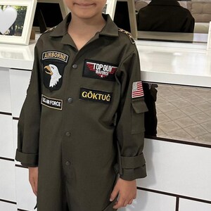 May include: A young person wearing an olive green flight suit and matching cap. The suit features patches that read "AIRBORNE", "U.S. AIR FORCE", "TOP GUN MAVERICK", and "G&Ouml;KTUĞ", along with an American flag patch.