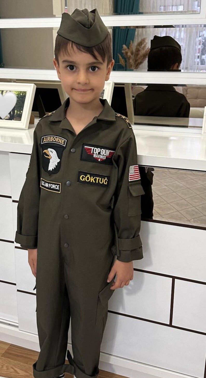 May include: A young person wearing an olive green flight suit and matching cap. The suit features patches that read "AIRBORNE", "U.S. AIR FORCE", "TOP GUN MAVERICK", and "G&Ouml;KTUĞ", along with an American flag patch.