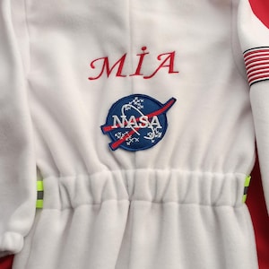 Op de afbeelding: Wit astronaut pak met een NASA-patch en de naam "MIA" geborduurd op de voorkant. Het pak heeft een capuchon en een trekkoord in de taille.