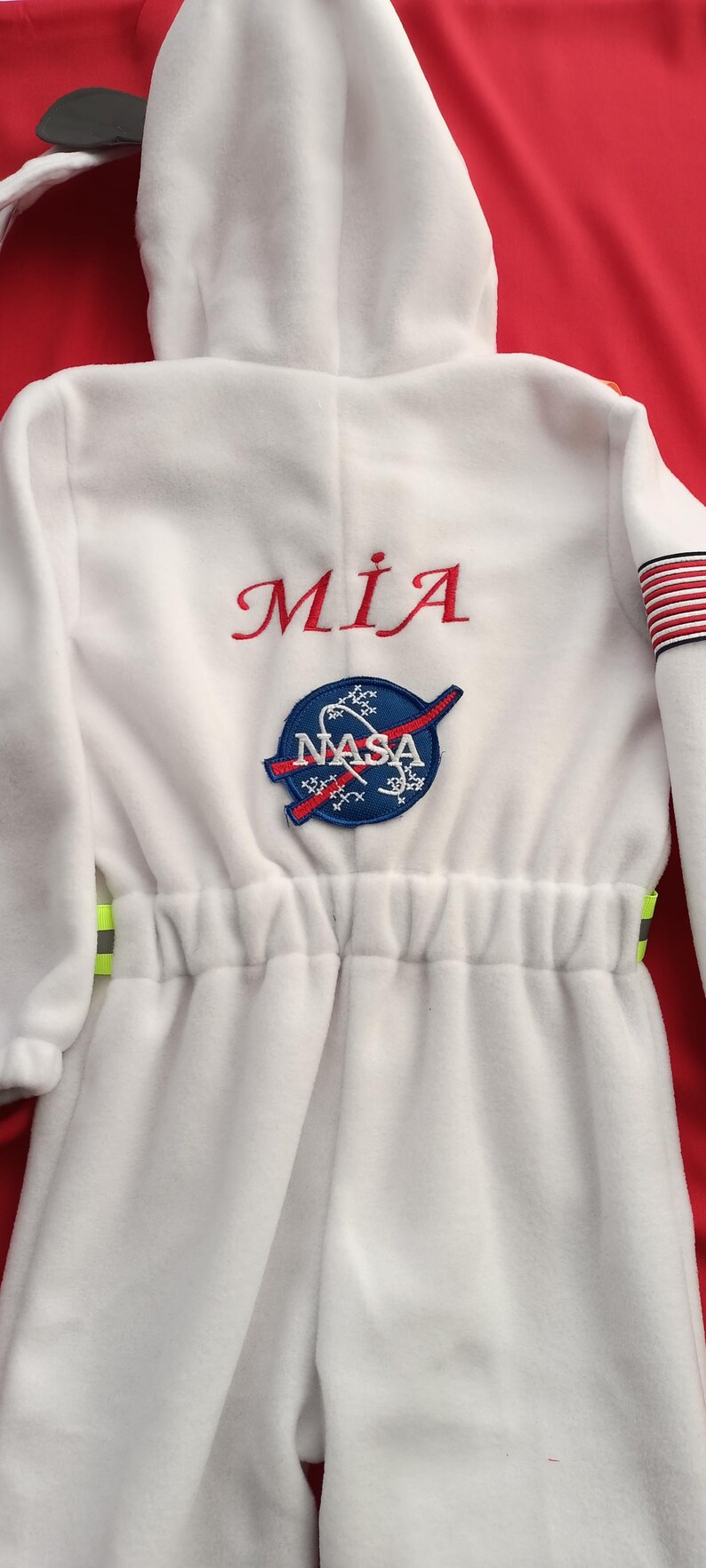 Op de afbeelding: Wit astronaut pak met een NASA-patch en de naam "MIA" geborduurd op de voorkant. Het pak heeft een capuchon en een trekkoord in de taille.