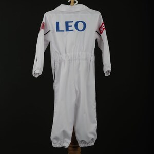 Może przedstawiać: Biały kombinezon astronauty z napisem "LEO" w niebieskich literach na plecach. Kombinezon ma długie rękawy, kołnierz oraz elastyczne mankiety i kostki. Kombinezon jest wystawiony na drewnianym stojaku.