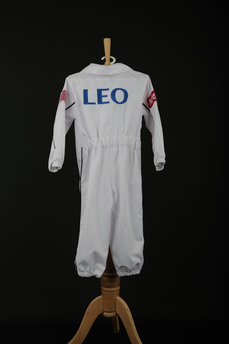 Może przedstawiać: Biały kombinezon astronauty z napisem "LEO" w niebieskich literach na plecach. Kombinezon ma długie rękawy, kołnierz oraz elastyczne mankiety i kostki. Kombinezon jest wystawiony na drewnianym stojaku.