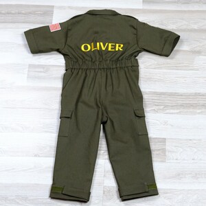 May include: Olive green children's jumpsuit with short sleeves, pockets, and the name "OLIVER" embroidered in yellow on the back. An American flag patch is on the left sleeve. The jumpsuit has an elastic waist and is made of a durable fabric.