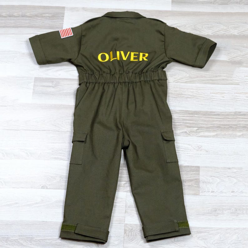 May include: Olive green children's jumpsuit with short sleeves, pockets, and the name "OLIVER" embroidered in yellow on the back. An American flag patch is on the left sleeve. The jumpsuit has an elastic waist and is made of a durable fabric.