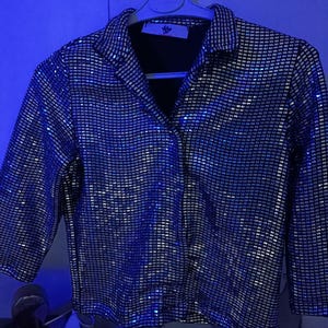 May include: A silver sequin button-down shirt with a black collar and cuffs. The shirt is hanging on a white hanger. The sequins create a shimmering effect, reflecting light. The shirt has a classic collared neckline and long sleeves.