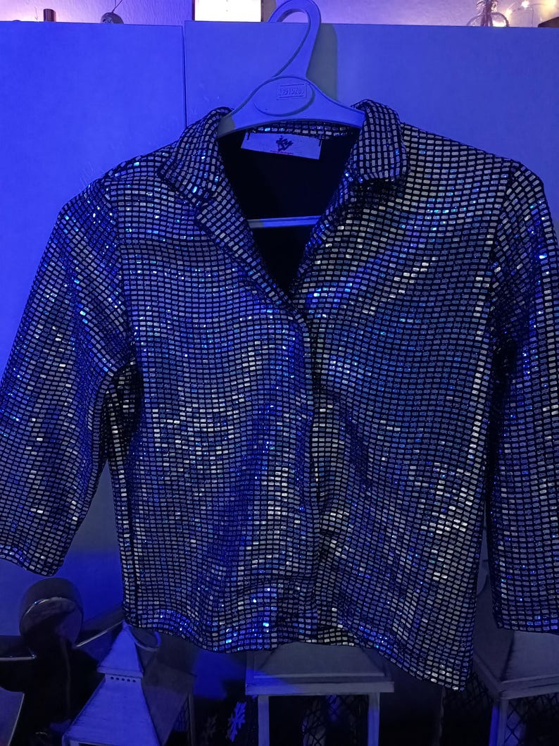 May include: A silver sequin button-down shirt with a black collar and cuffs. The shirt is hanging on a white hanger. The sequins create a shimmering effect, reflecting light. The shirt has a classic collared neckline and long sleeves.
