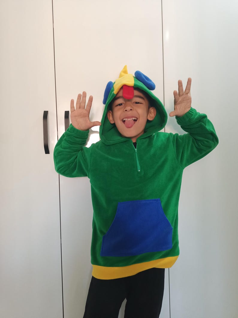 Puede incluir: Un ni&ntilde;o con una sudadera con capucha verde, azul, amarilla y roja con un dise&ntilde;o de p&aacute;jaro colorido en la capucha. El ni&ntilde;o sonr&iacute;e y saca la lengua.
