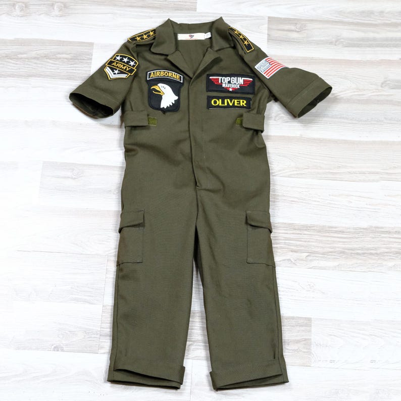May include: Olive green jumpsuit with short sleeves, multiple pockets, and patches. Patches include "Army", "Airborne", a bald eagle, a "Top Gun Maverick" logo, and the name "Oliver". The jumpsuit has a collar and a US flag patch.