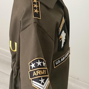 Puede incluir: Disfraz de estilo militar verde oliva con parches dorados y negros. Los parches incluyen el texto "ARMY" y "U.S. AIR FORCE", junto con dise&ntilde;os de estrellas y galones. El disfraz tiene cuello y cintura entallada.