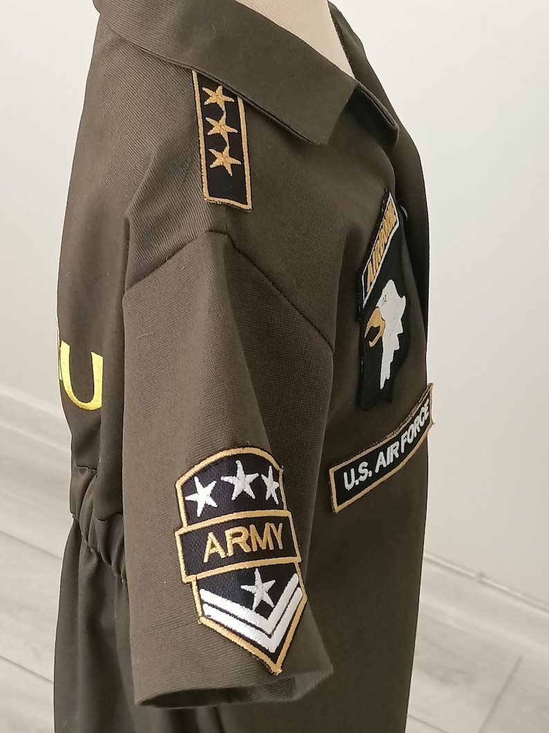 Puede incluir: Disfraz de estilo militar verde oliva con parches dorados y negros. Los parches incluyen el texto "ARMY" y "U.S. AIR FORCE", junto con dise&ntilde;os de estrellas y galones. El disfraz tiene cuello y cintura entallada.