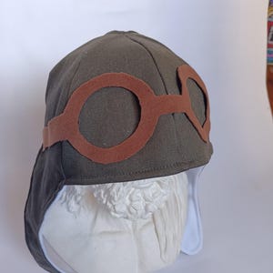 May include: An olive green aviator hat with brown felt goggles. The hat has a rounded crown and ear flaps with a white lining. The goggles are attached to the front of the hat with a brown band. The hat is displayed on a white bust.