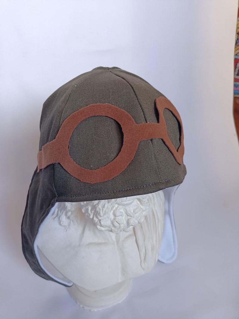 May include: An olive green aviator hat with brown felt goggles. The hat has a rounded crown and ear flaps with a white lining. The goggles are attached to the front of the hat with a brown band. The hat is displayed on a white bust.