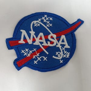 Może przedstawiać: Haftowana naszywka NASA z niebieskim okrągłym wzorem, białym logo NASA, czerwoną smugą i białymi akcentami w kształcie gwiazd. Naszywka znajduje się na białym tle materiału.