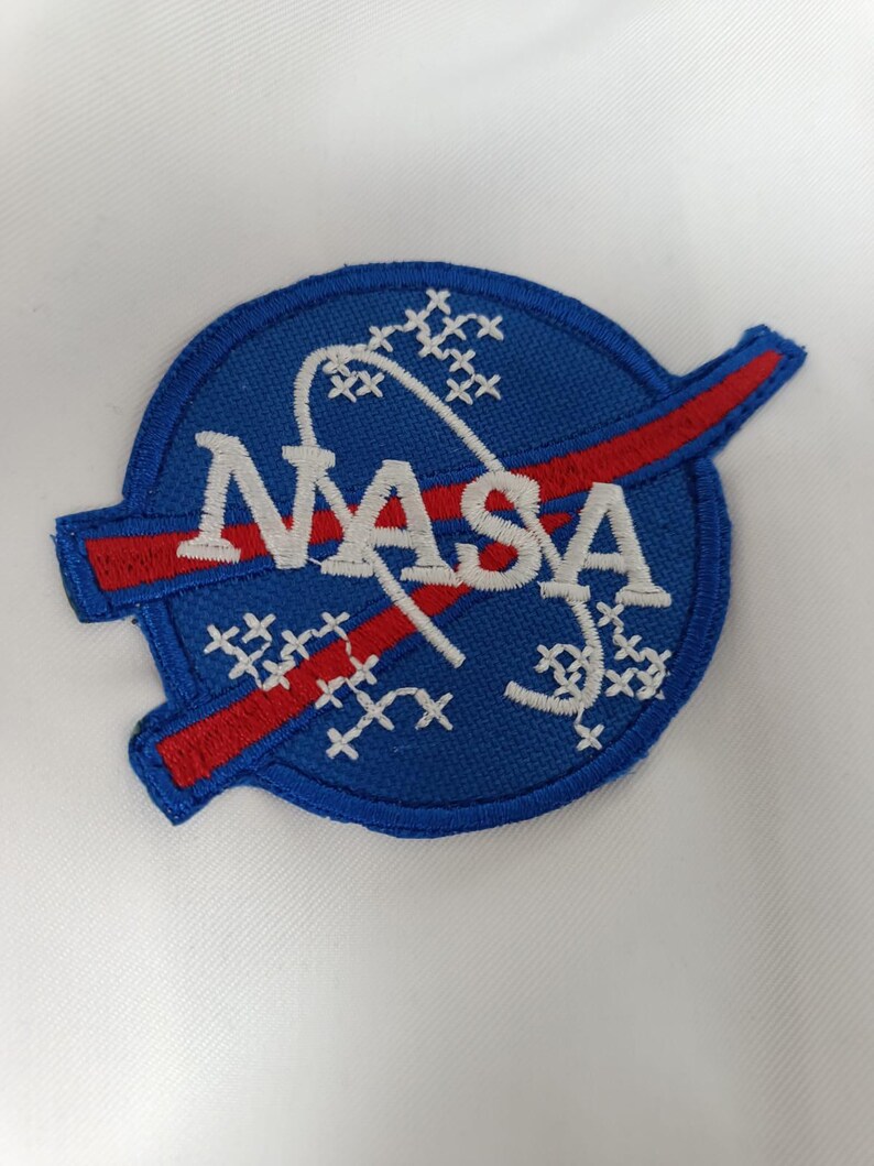 Może przedstawiać: Haftowana naszywka NASA z niebieskim okrągłym wzorem, białym logo NASA, czerwoną smugą i białymi akcentami w kształcie gwiazd. Naszywka znajduje się na białym tle materiału.