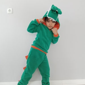 Peut inclure: Un enfant portant un costume de dinosaure vert et orange avec une t&ecirc;te de dinosaure blanche et verte. Le costume a une capuche et une queue.
