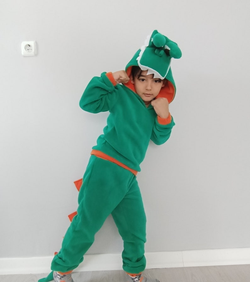 Peut inclure: Un enfant portant un costume de dinosaure vert et orange avec une t&ecirc;te de dinosaure blanche et verte. Le costume a une capuche et une queue.