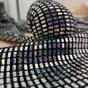 May include: Close-up of a black fabric covered in small, rectangular, iridescent sequins. The sequins reflect a range of colors, including silver, blue, and purple. The fabric is likely used for clothing or accessories.