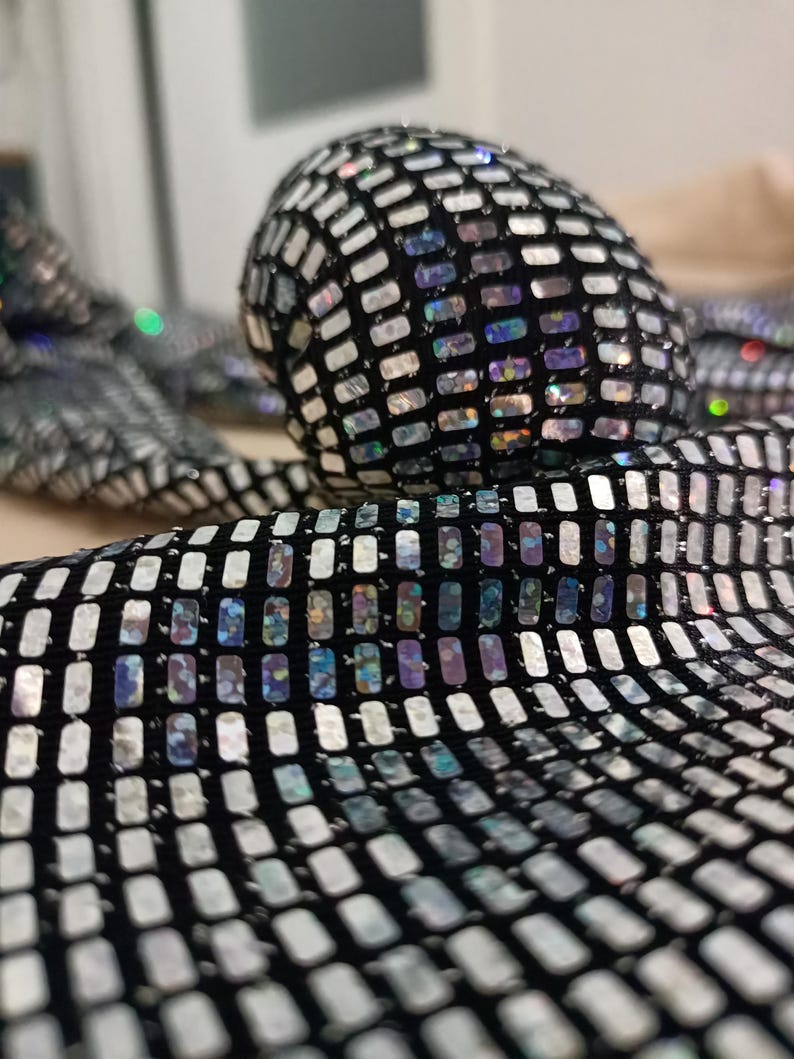 May include: Close-up of a black fabric covered in small, rectangular, iridescent sequins. The sequins reflect a range of colors, including silver, blue, and purple. The fabric is likely used for clothing or accessories.